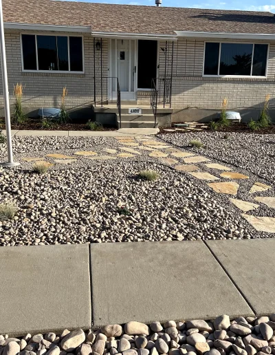 Gravel‑covered front yard with scattered plants, concrete walkway, and a brick house.