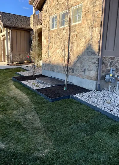 Neatly edged garden beds with young trees and decorative rocks beside a stone house.